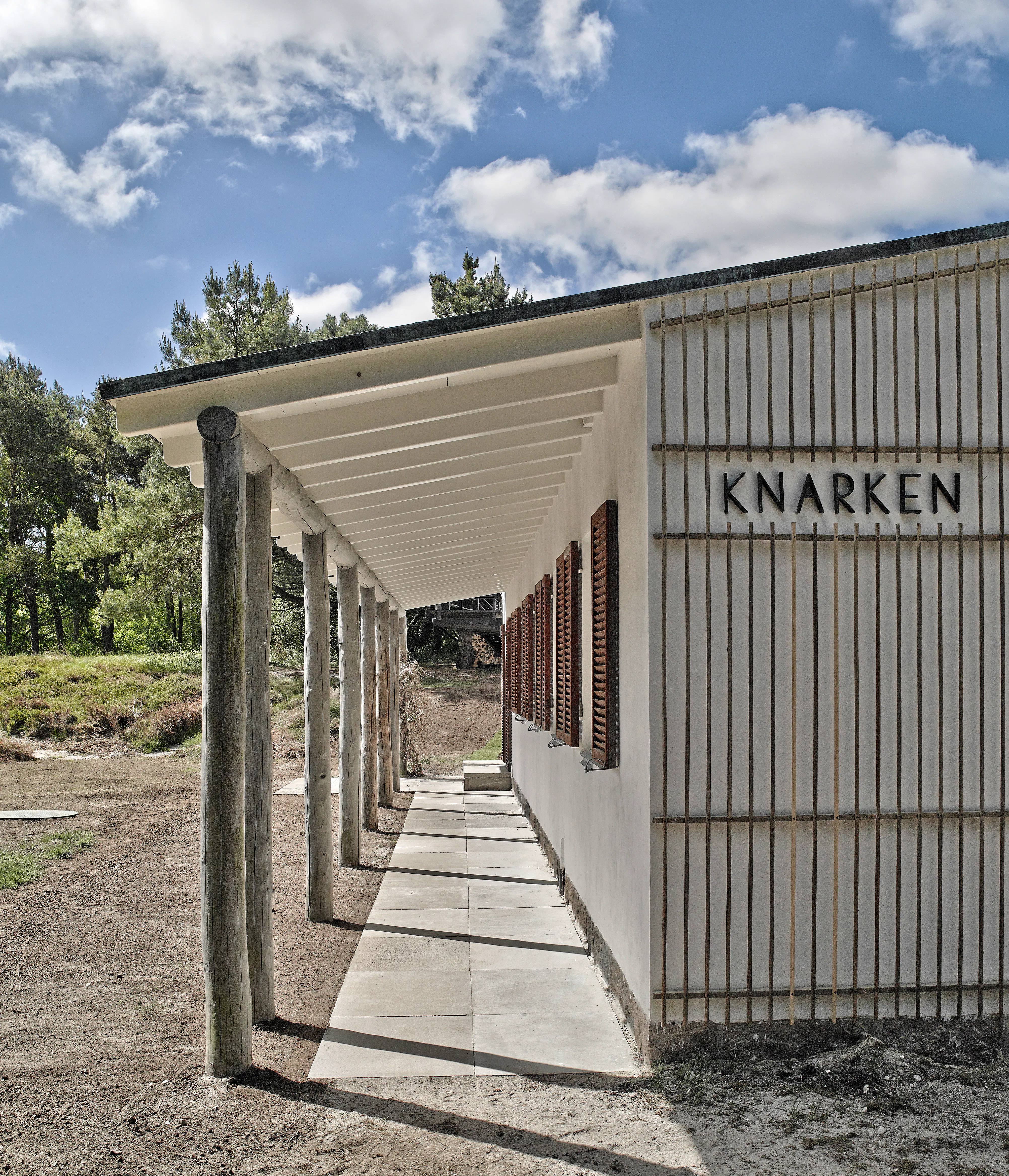 Side af garage til Arne Jacobsens sommerhus med overdækket gang, træstolper, hvide vægge og skoddevinduer.