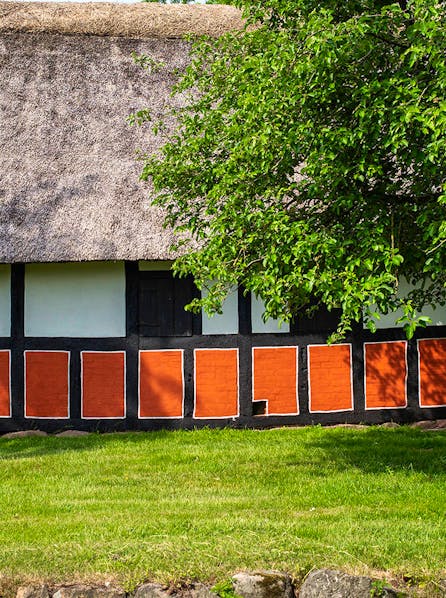 Bindingsværksgård med stråtag set fra siden med stort træ i højre side af billedet.