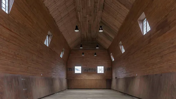 Tom ridehal med højt loft, træbeklædte vægge og små vinduer, oplyst af sorte loftslamper.