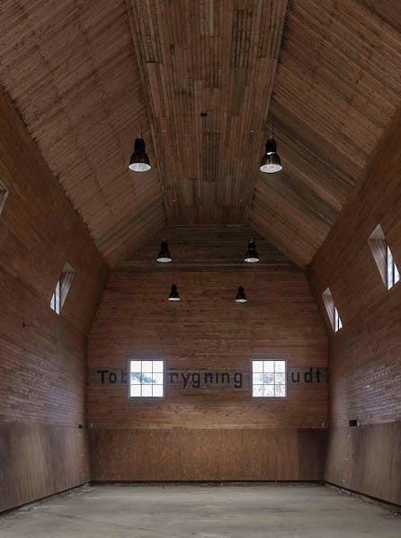 Tom ridehal med højt loft, træbeklædte vægge og små vinduer, oplyst af sorte loftslamper.