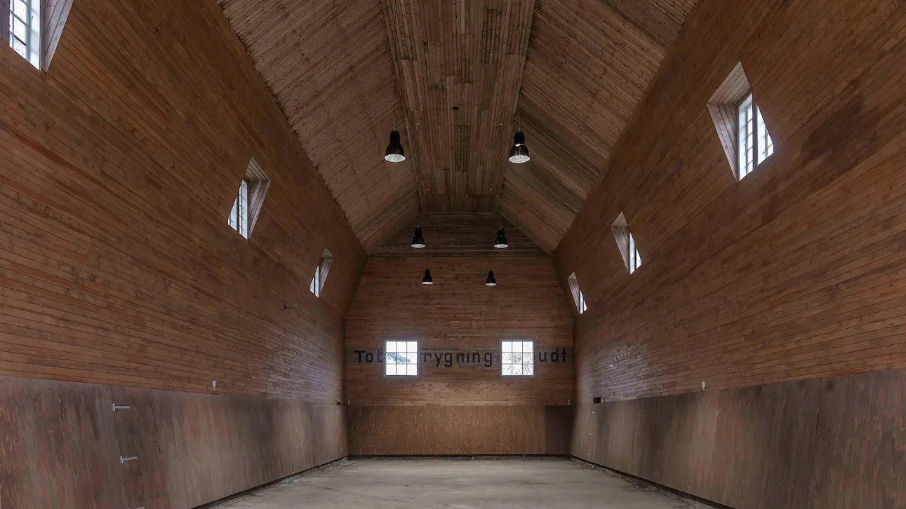 Tom ridehal med højt loft, træbeklædte vægge og små vinduer, oplyst af sorte loftslamper.