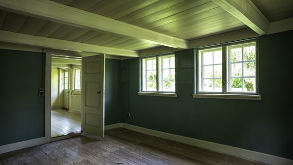 Mørkt rum med vinduer, grønne vægge, mørkt trægulv og hvidmalet træloft.