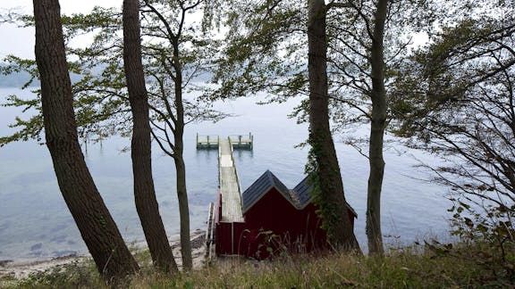 Badebro ved Hindsgavl Slot med udsigt over vandet og lille rødt badehus i skovklædt skråning.
