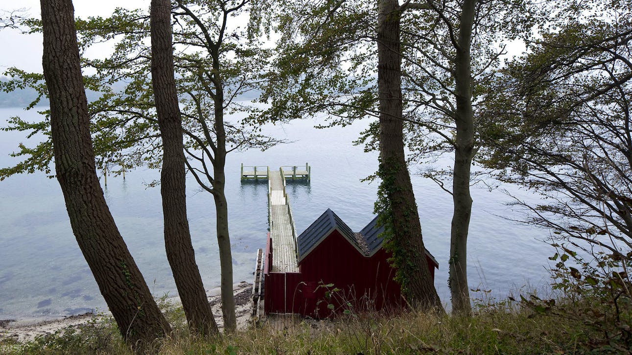 Badebro ved Hindsgavl Slot med udsigt over vandet og lille rødt badehus i skovklædt skråning.
