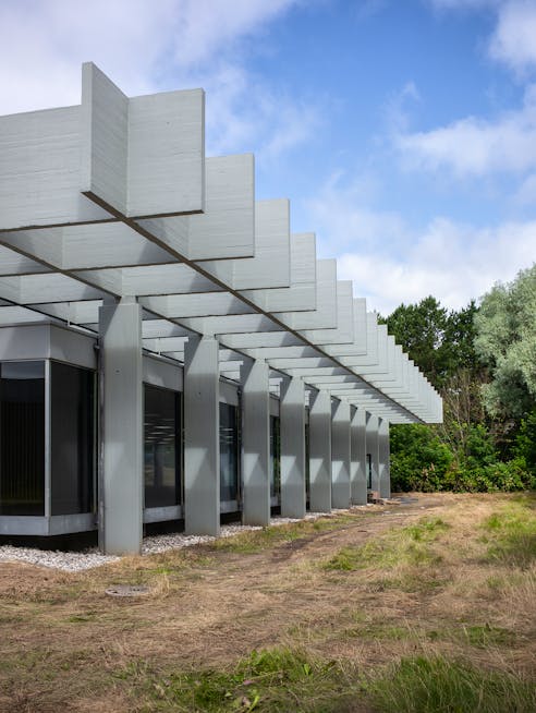 Bygning med søjler og horisontale betonelementer, omgivet af græs og træer under blå himmel.