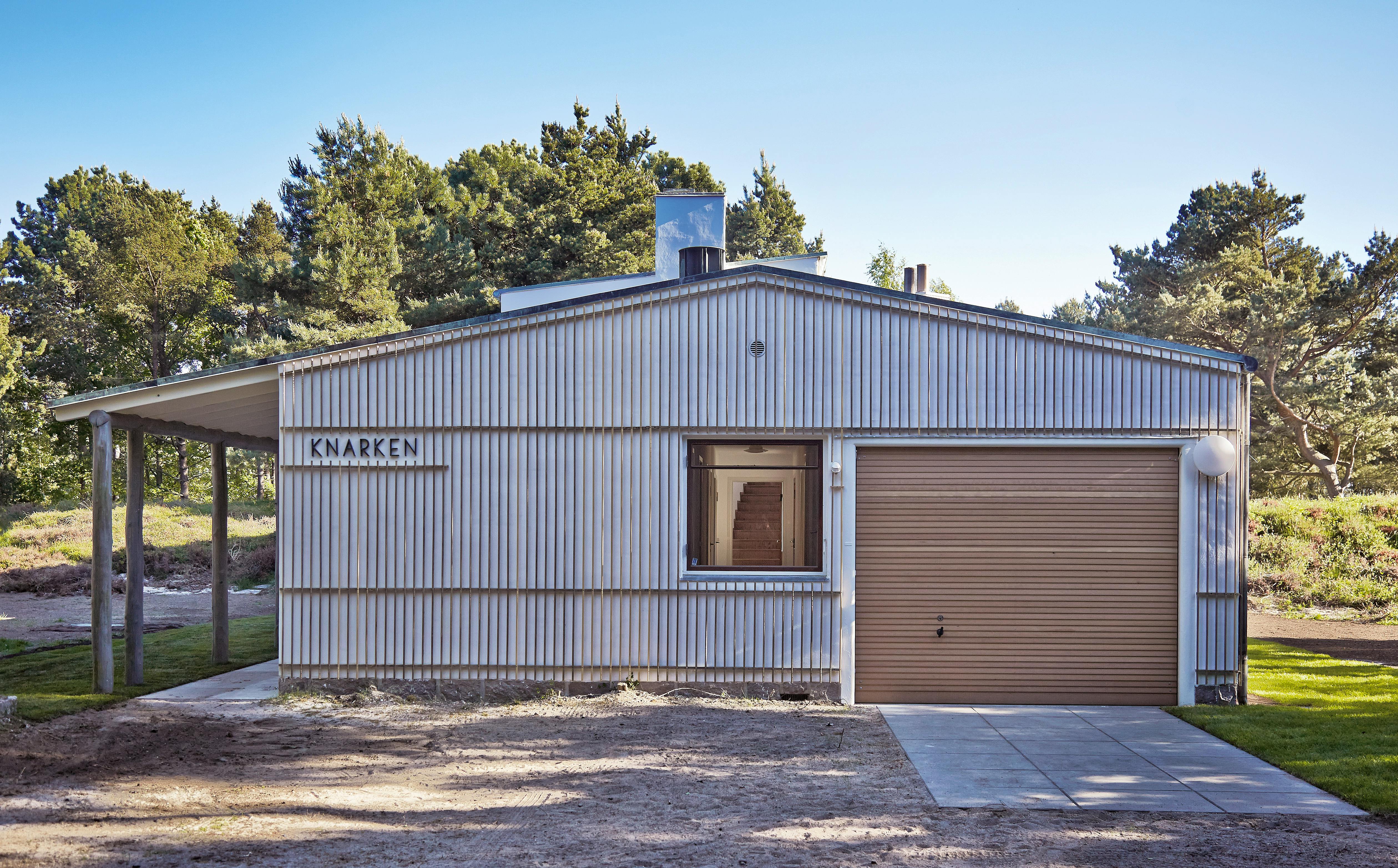 Garagen til Arne Jacobsens sommerhus. Rektangulær bygning med lodrette trælameller, skråt tag, vindue, rullegarageport og skiltet 'KNARKEN'.
