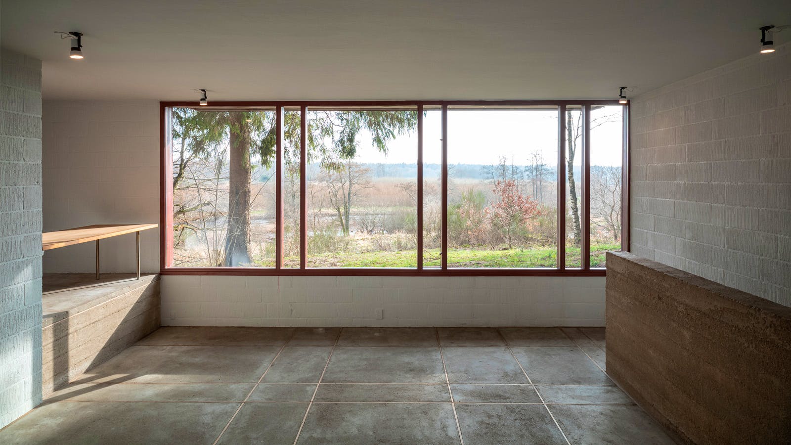 Stue i Poul Erik Thyrrings eget hus med panoramavindue mod naturen, vægge af kalket gasbeton, gulv af støbt beton og halvmur i rå in-situstøbt beton.