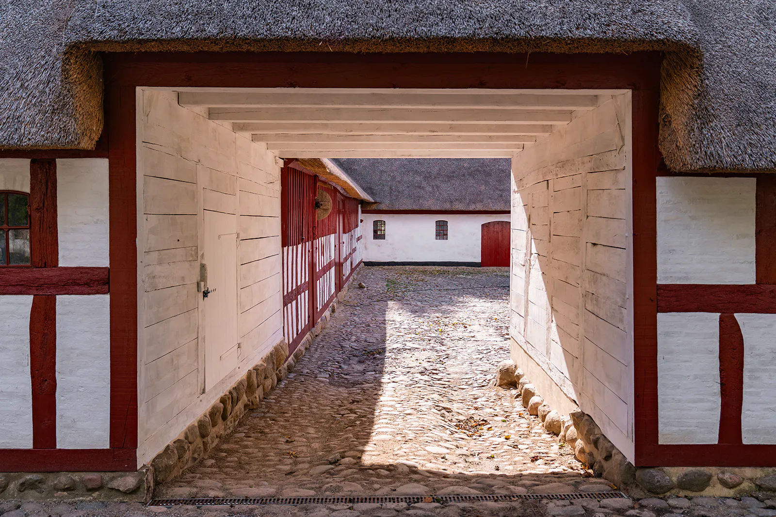 Porten til gårdspladsen – med kig ind til bindingsværkslænger, brostensbelægning og stråtækte tage.