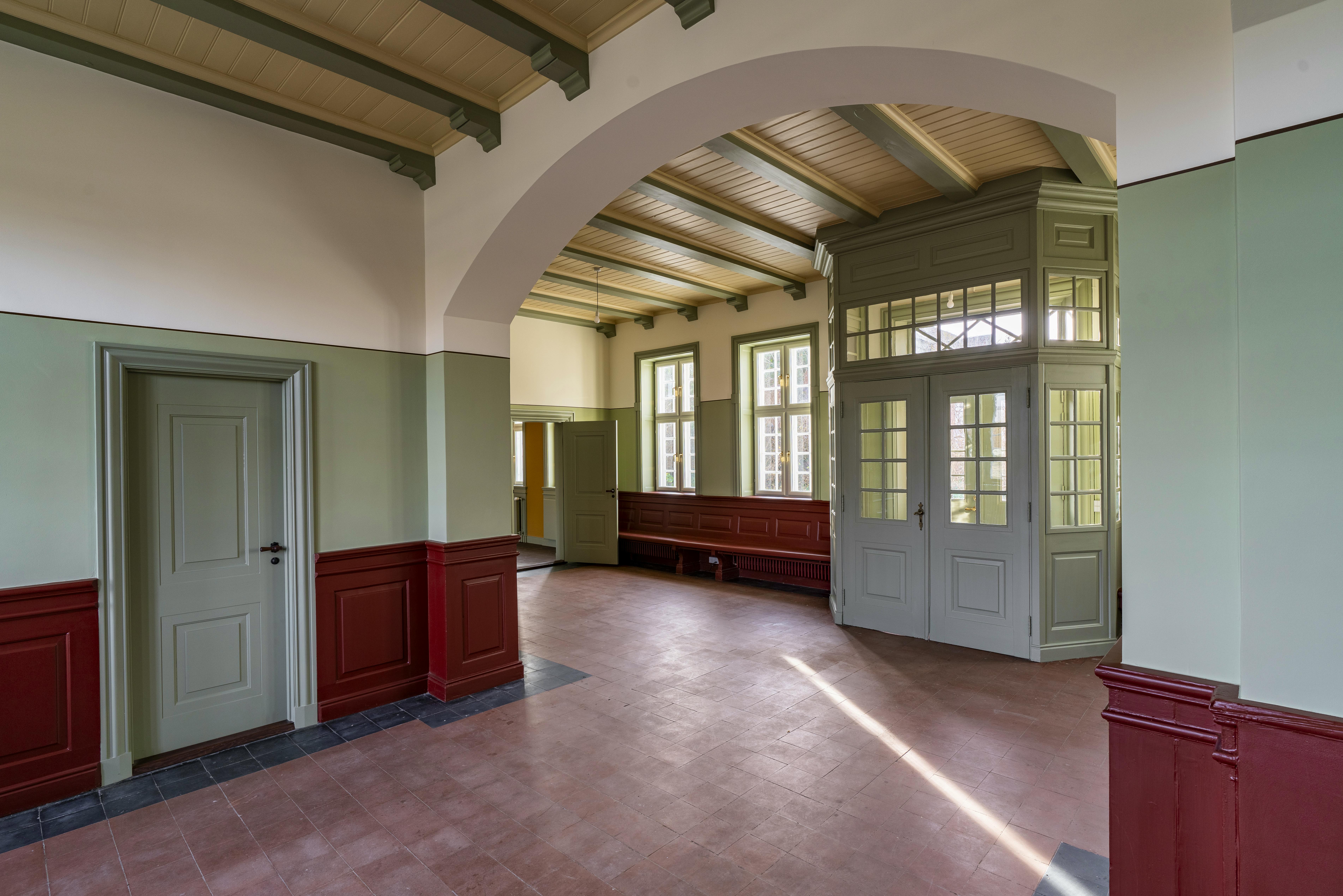 Fra døråbningen ses ventesalens vindfang i perspektiv, omgivet af grønne karme og røde paneler.