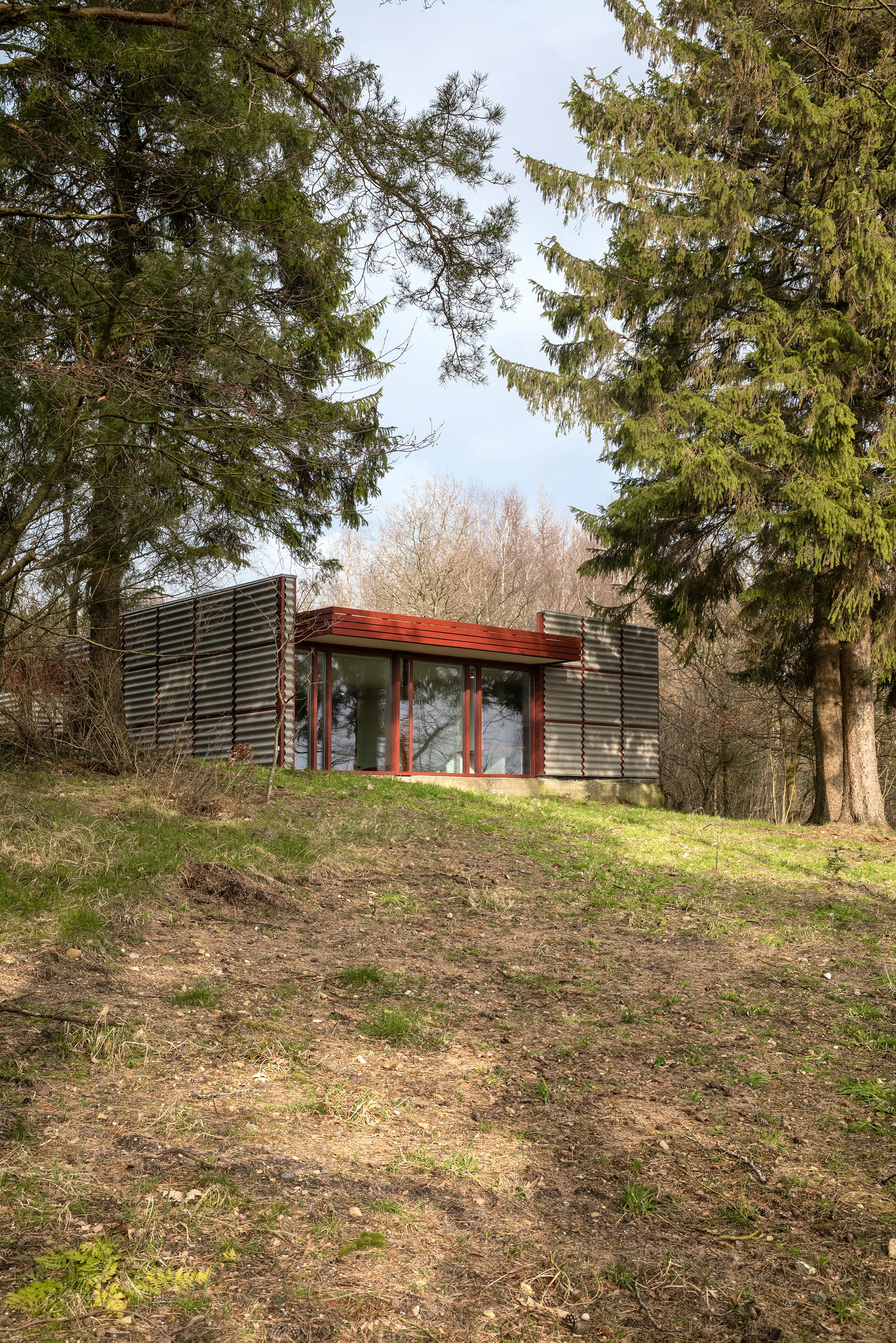 Poul Erik Thyrrings eget hus med bølgende metalfacade, røde vinduesrammer og fladt tag, beliggende på en skrånende naturgrund omgivet af høje træer.