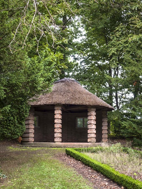 En lille, åben pavillon med søjler af mursten og stråtag står omgivet af tætte grønne træer. Foran pavillonen fører en græssti op til bygningen, og en lav, velplejet hæk løber langs højre side af stien.