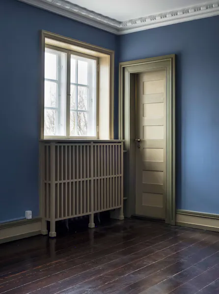 Et rum med mørkt trægulv, blå vægge og hvid stuk langs loftet, hvor et vindue med sprosser og en dør med felter er indrammet af olivengrønne karme, og en radiator skjules bag en tremmebeklædt kappe under vinduet.