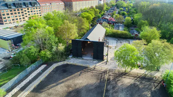 Dronefoto af Ballonhangaren som viser ridebanen, hangaren, et beboelsesområde og grønne træer rundt om hangaren.