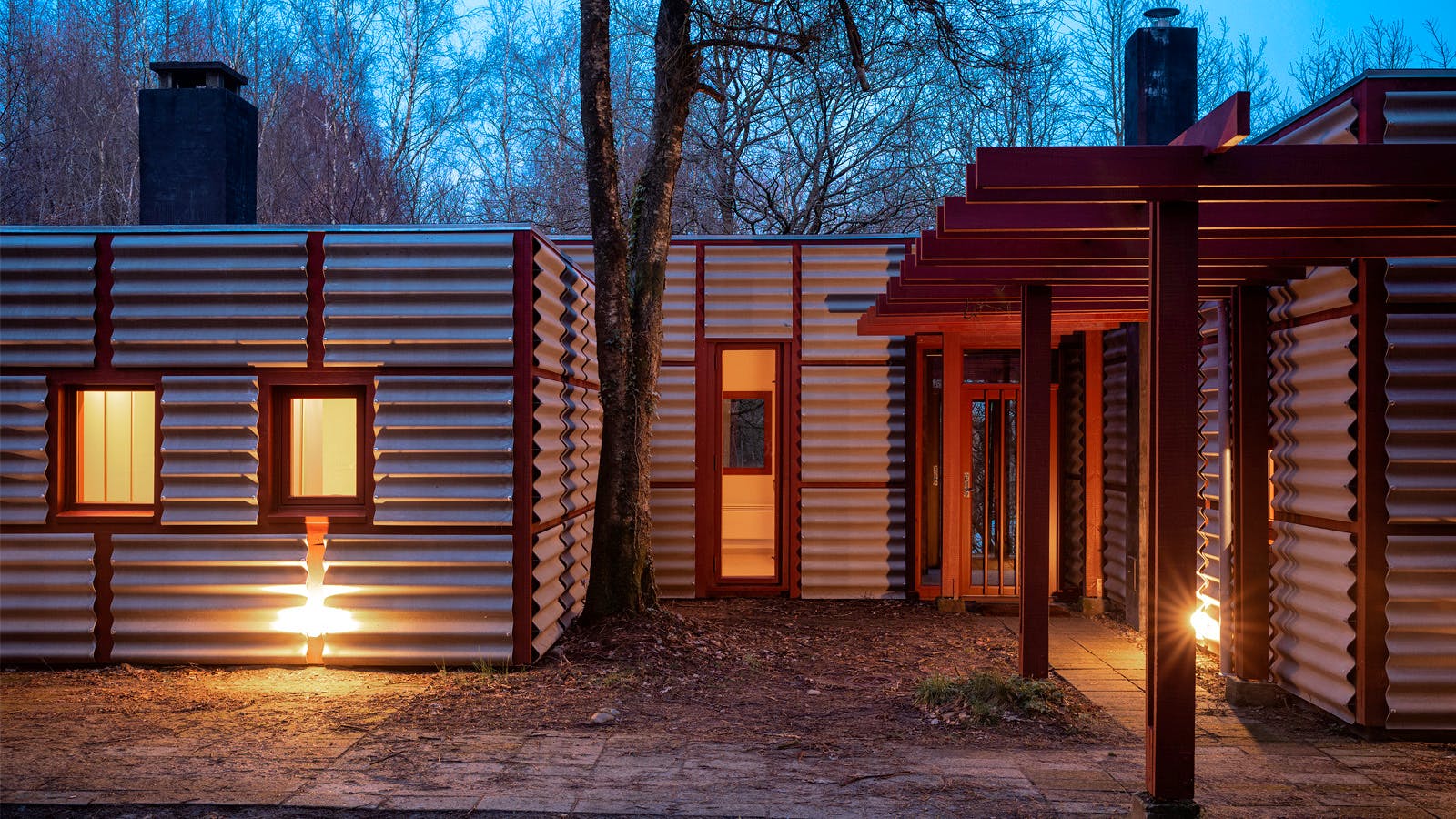 Poul Erik Thyrrings eget hus i skumring med bølget metalfacade, rød pergola og varmt lys indefra og langs facaden.