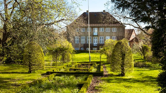En veltrimmet have med formklippede buske og symmetriske bede fører op til en stor murstensbygning med rødt tegltag og hvide vinduer, omgivet af frodige træer og forårsgrønt.