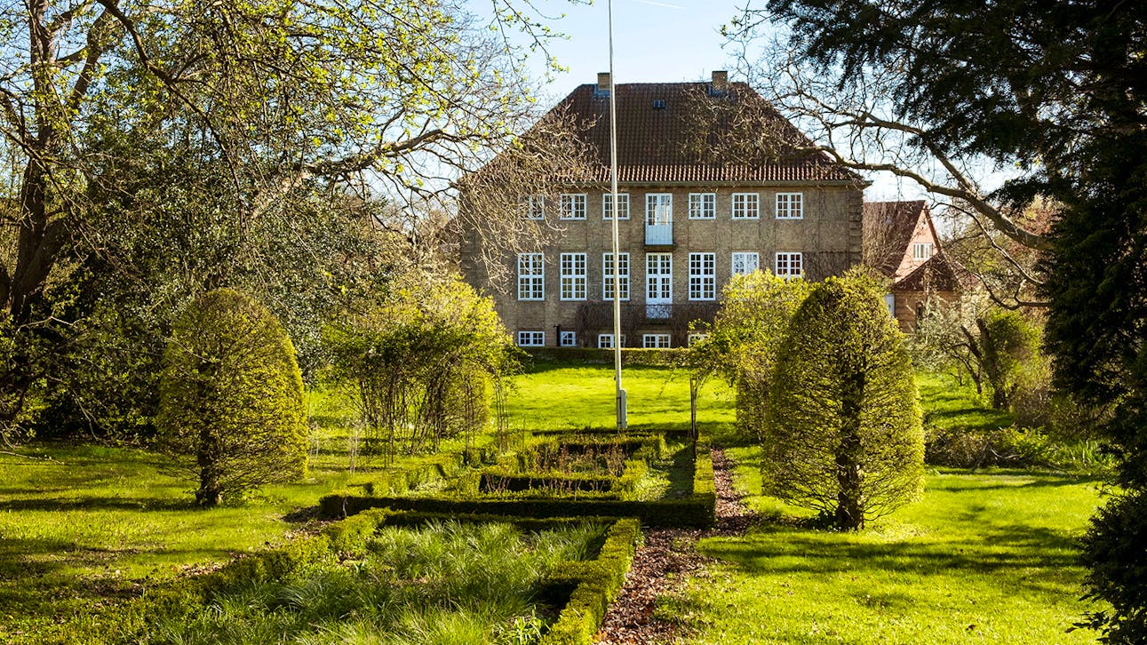 En veltrimmet have med formklippede buske og symmetriske bede fører op til en stor murstensbygning med rødt tegltag og hvide vinduer, omgivet af frodige træer og forårsgrønt.