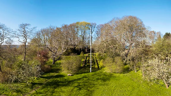 En velplejet græsplæne med symmetriske hække og stier fører op til en pavillon, omgivet af løv- og frugttræer i tidligt forår under en klar blå himmel.