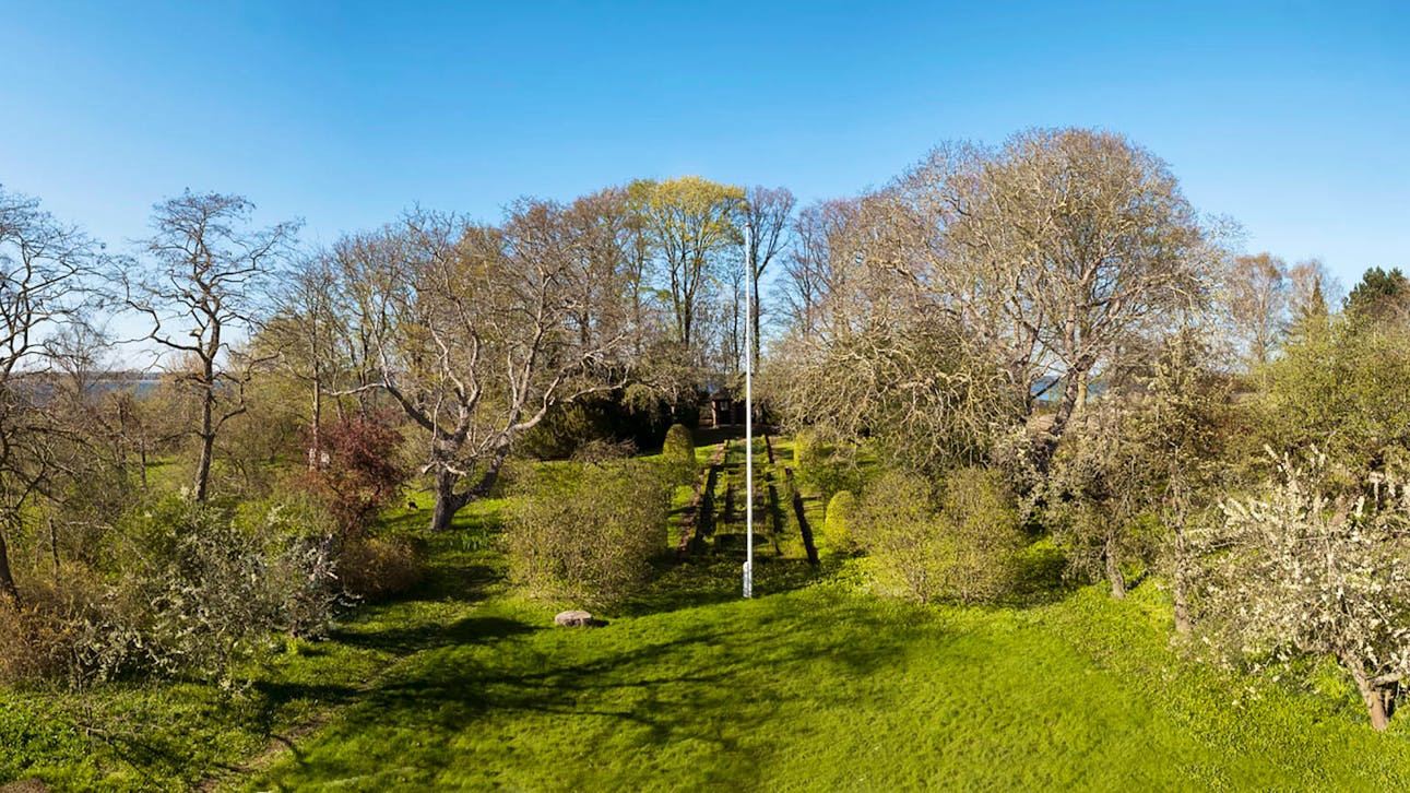 En velplejet græsplæne med symmetriske hække og stier fører op til en pavillon, omgivet af løv- og frugttræer i tidligt forår under en klar blå himmel.