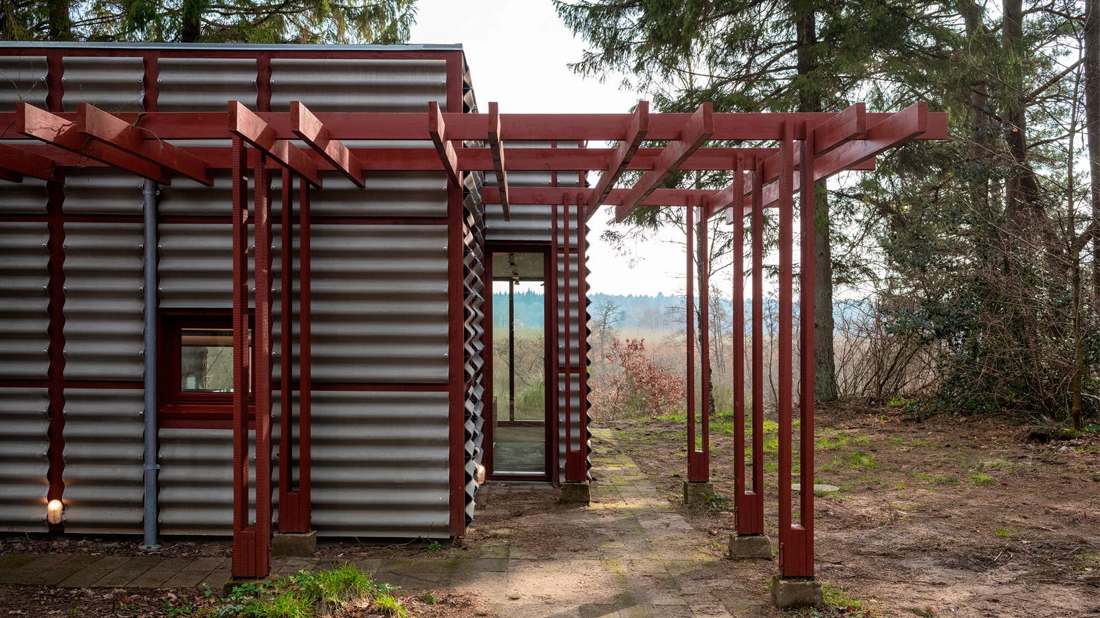 Rød pergola foran facade med bølgede metalplader og rødmalede vinduesrammer ved Poul Erik Thyrrings eget hus, omgivet af skov og natur.