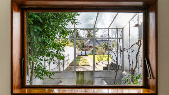 Udsigt gennem vindue fra tegnestuen til et lille drivhus med glasloft og videre ud til haven.