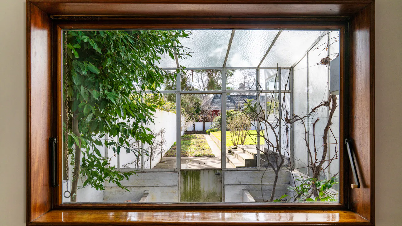 Udsigt gennem vindue fra tegnestuen til et lille drivhus med glasloft og videre ud til haven.