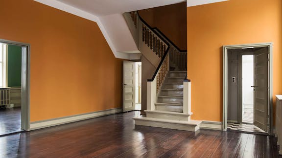 Et interiørbillede af en entré eller hall med mørkt trægulv, orange vægge, en hvidmalet trappe med mørke gelændere og adgang til flere rum gennem døre i grålige nuancer.