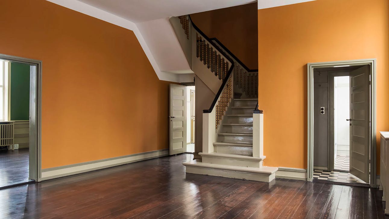 Et interiørbillede af en entré eller hall med mørkt trægulv, orange vægge, en hvidmalet trappe med mørke gelændere og adgang til flere rum gennem døre i grålige nuancer.