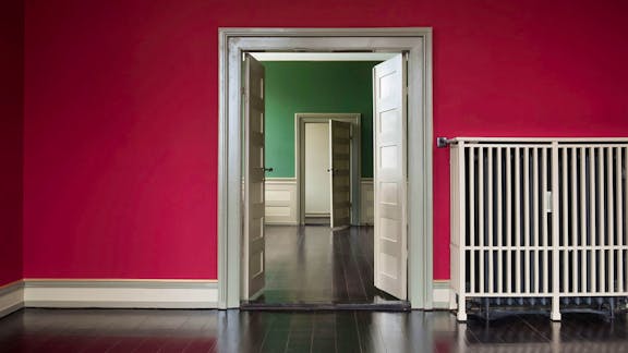 Et interiørbillede med en stærkt rødmalet væg, en hvidmalet radiator med tremmer foran, og et kig gennem flere åbne døre ind i rum med grønne vægge og mørkt trægulv.