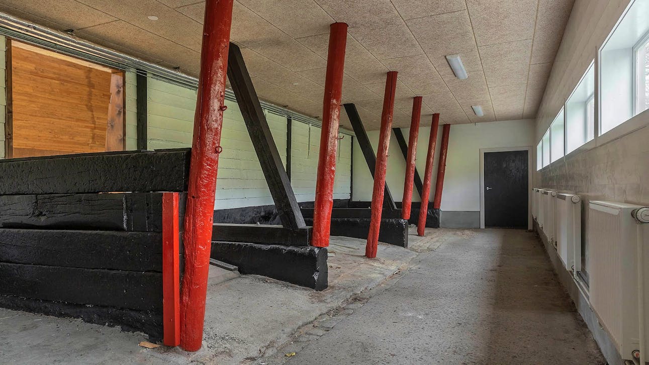 Stald med røde bjælker som står i stærk kontrast til de sorte vægge og murværk, som de står ved og på, samt betongulv og loftplader af træbeton.