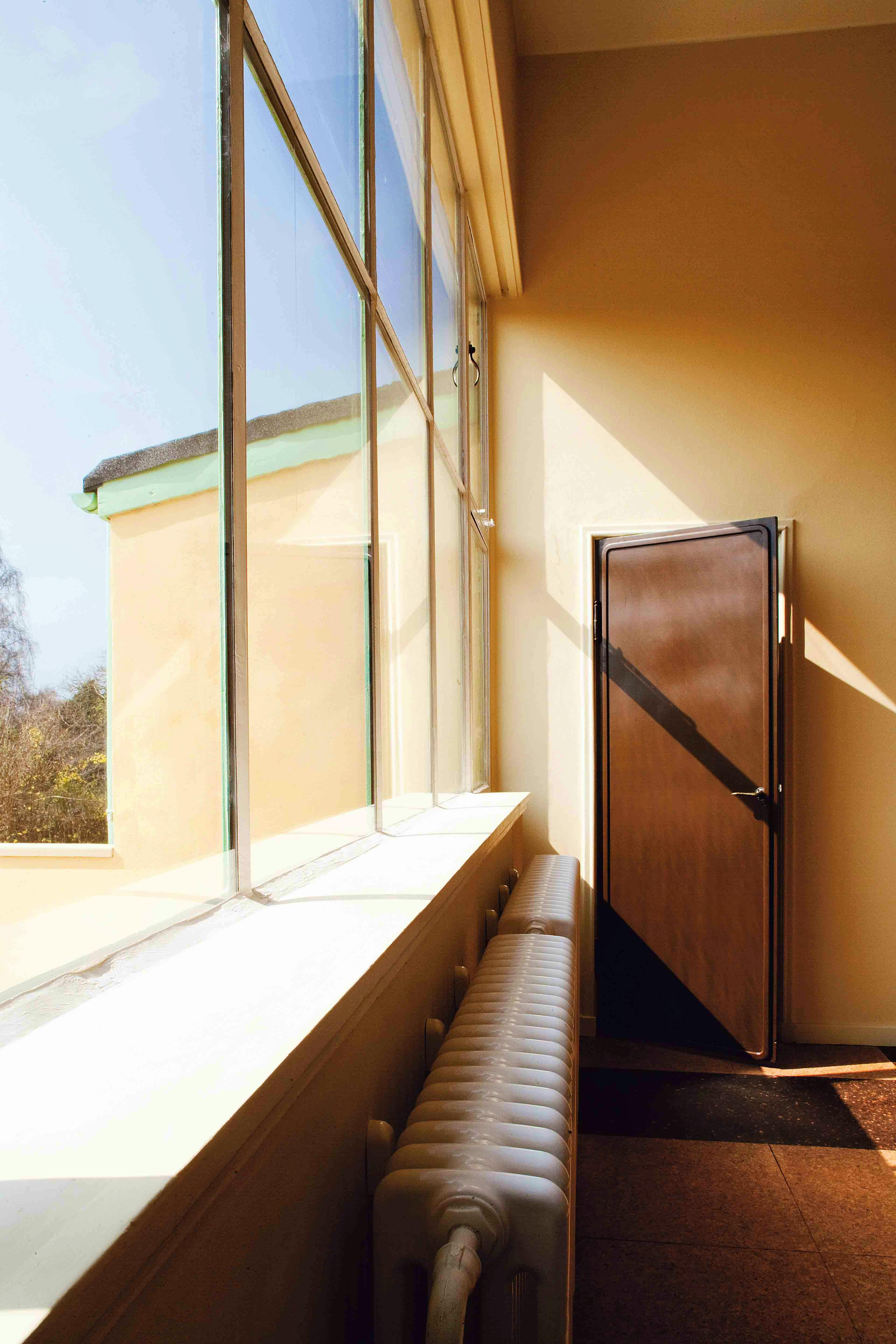 Lyst rum med stort stuevindue, hvid radiator og brun dør til tegnestuen