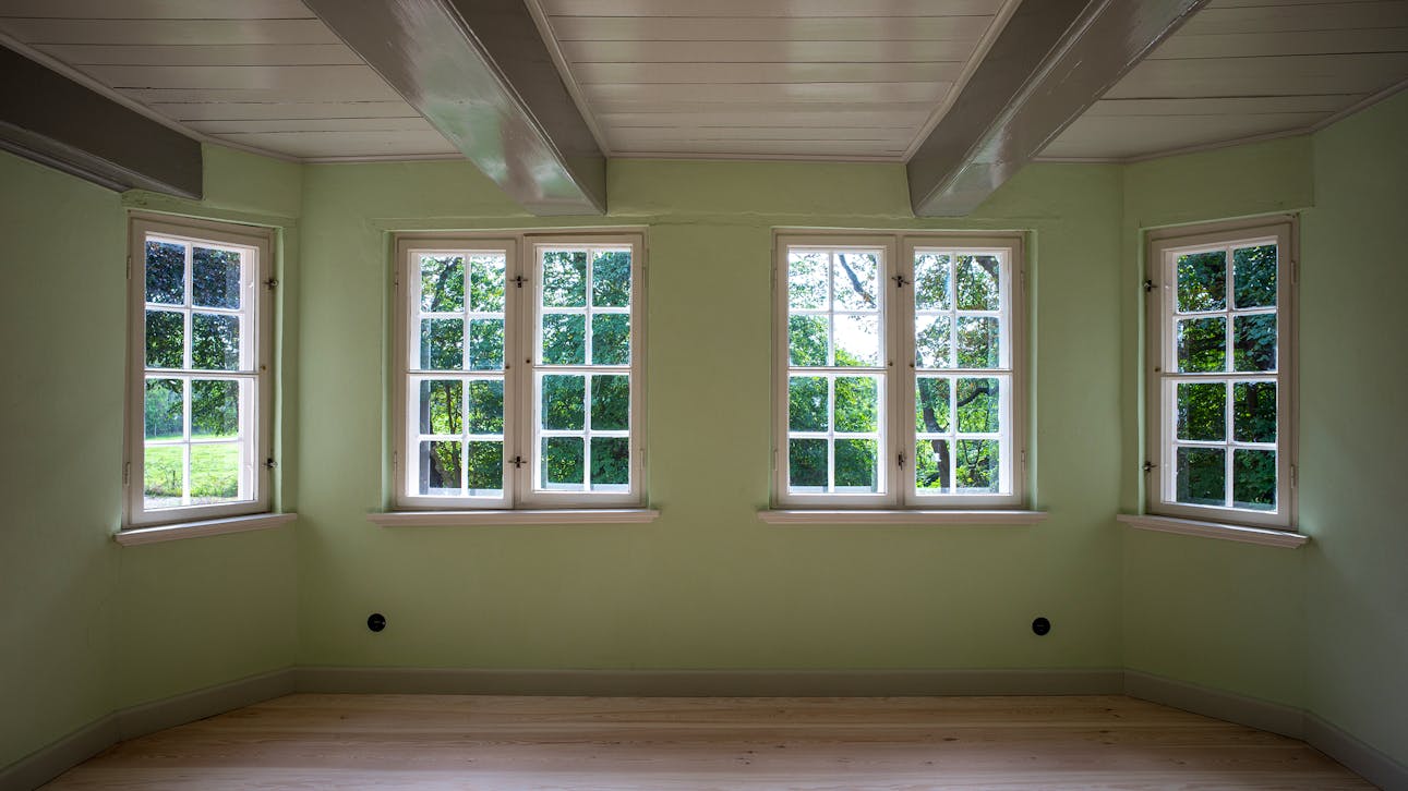 Lys grøn stue på Nørre Sødam med karnap, fire vinduer, lyse trægulve og synlige loftbjælker.