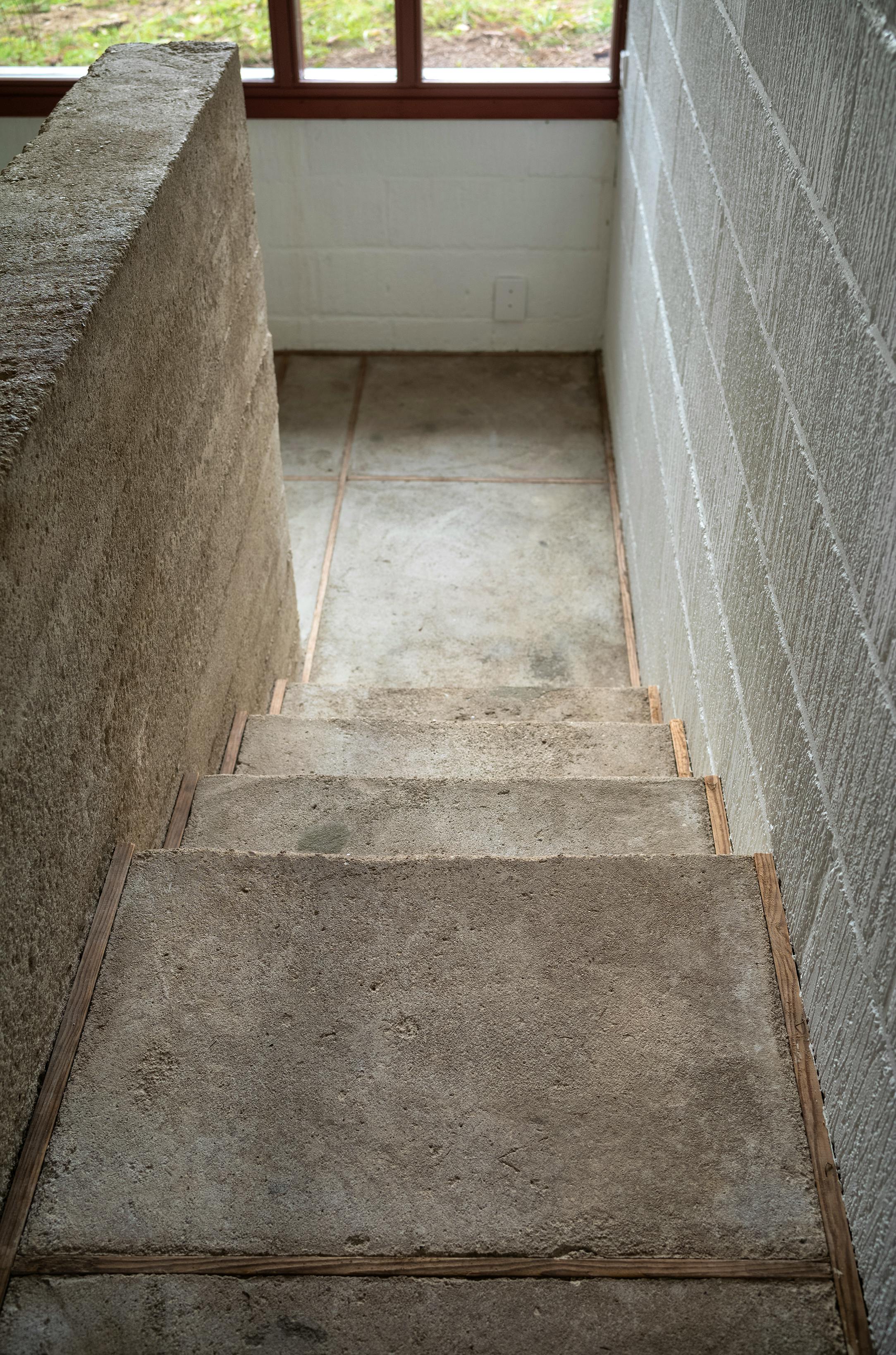 Trappe med rå betontrin og vægge fører ned mod et gulv med store klinker og et vindue ud mod have.
