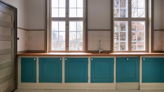 Et lyst og charmerende køkken med store sprossevinduer, blågrønne skabe med hvide greb og en varm træbordplade, der sammen skaber et enkelt og funktionelt udtryk.