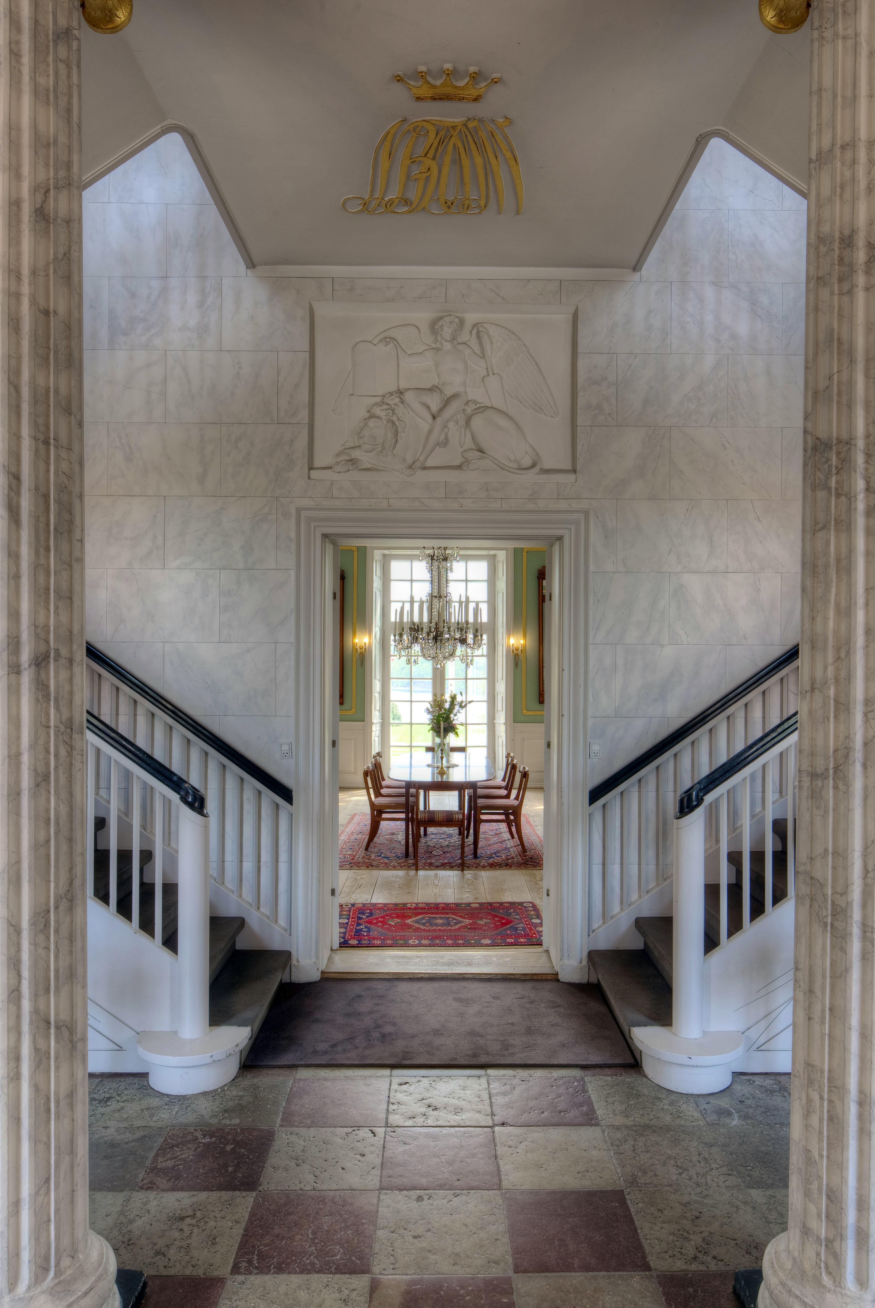 Hall på Hindsgavl Slot med trapper, søjler og kig ind til lyse stuer med lysekrone.