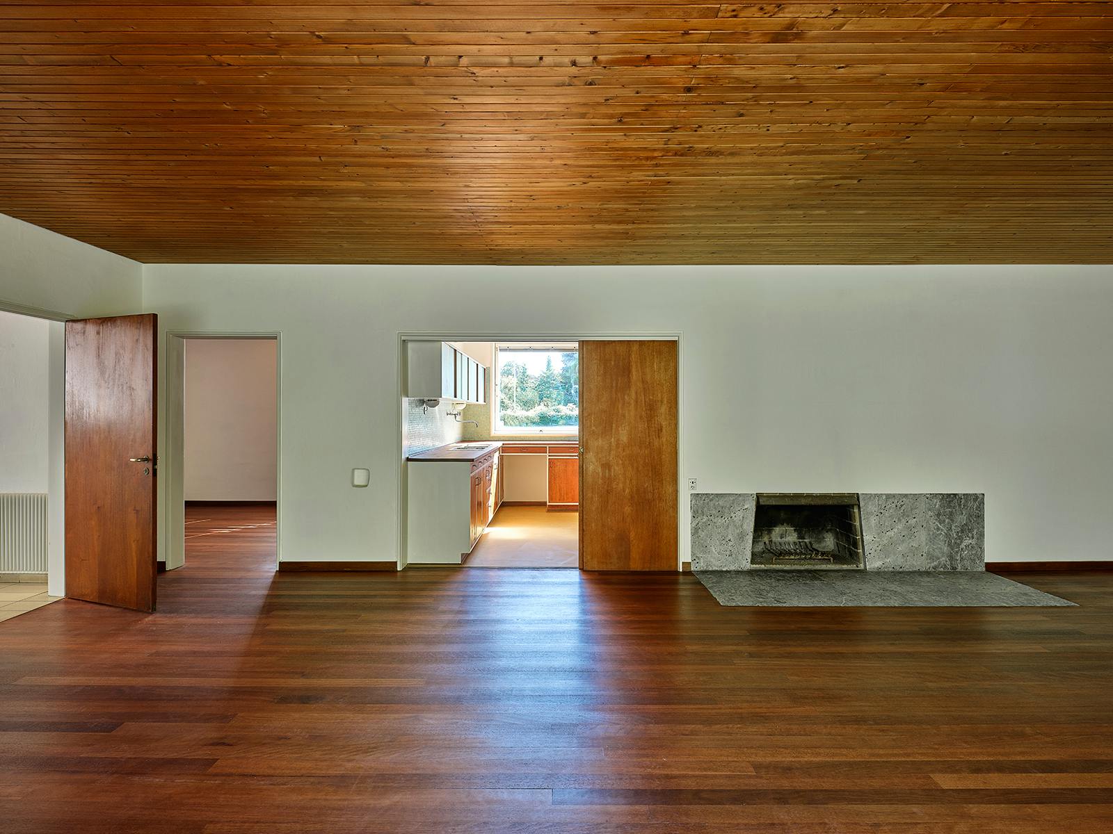 Stue i Vilhelm Lauritzens eget hus – åbent rumforløb med lav pejs i grønlandsk marmor, varme træflader og kig til det integrerede køkken.