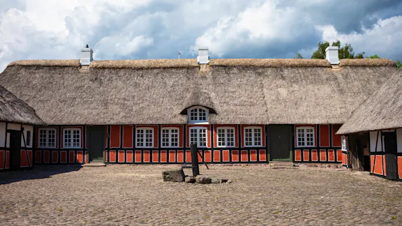 Bindingsværksgård med stråtag og brostensbelagt gårdsplads under dramatiske skyer.