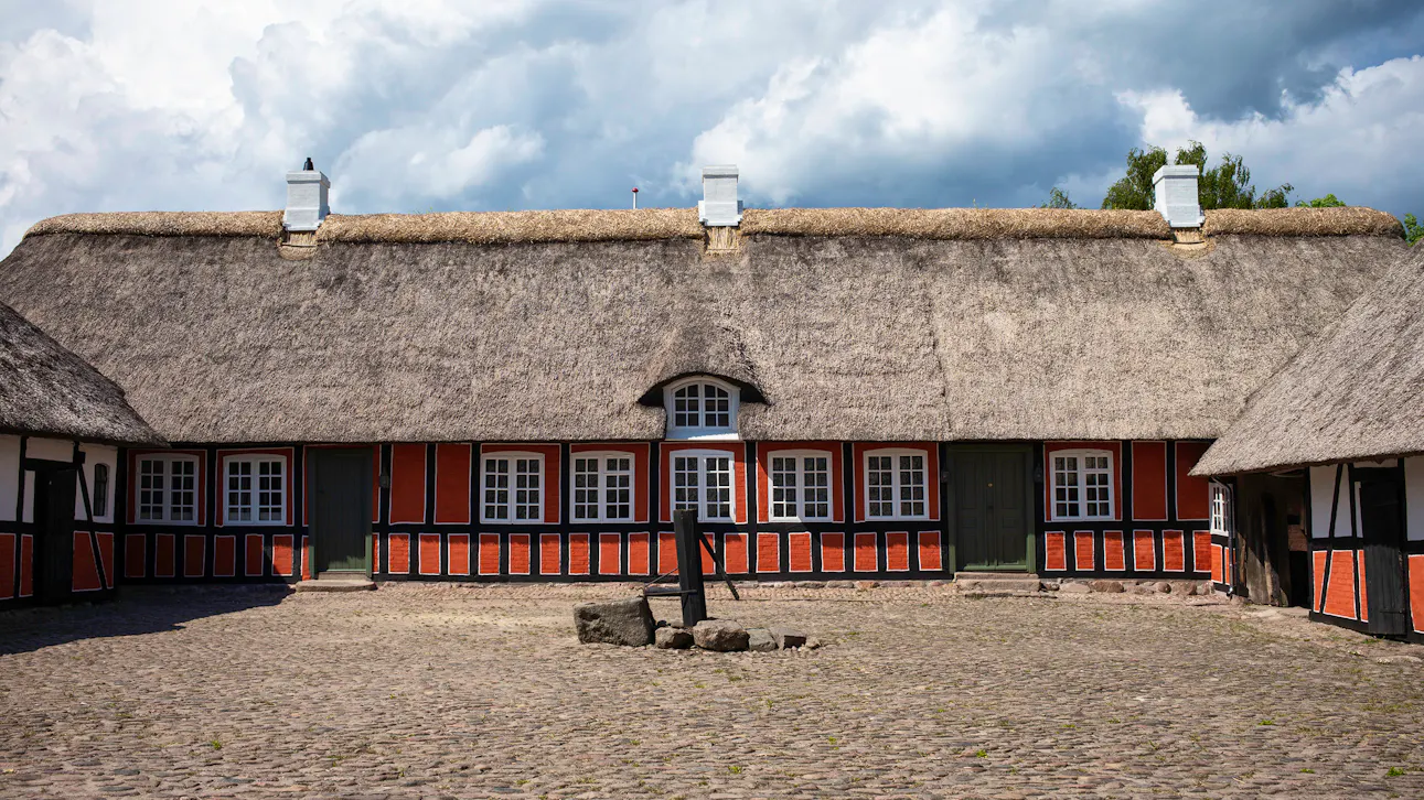 Bindingsværksgård med stråtag og brostensbelagt gårdsplads under dramatiske skyer.