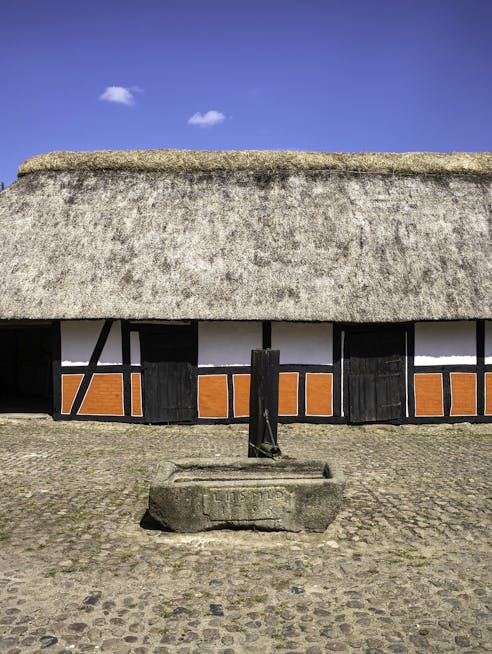 Brostensbelagt gårdsplads med vandpumpe i midten og bindingsværksgård i baggrunden under blå himmel.