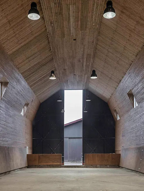 Tom ridehal med højt loft, træbeklædte vægge og små vinduer, oplyst af sorte loftslamper med kig mod portene og udgangen.