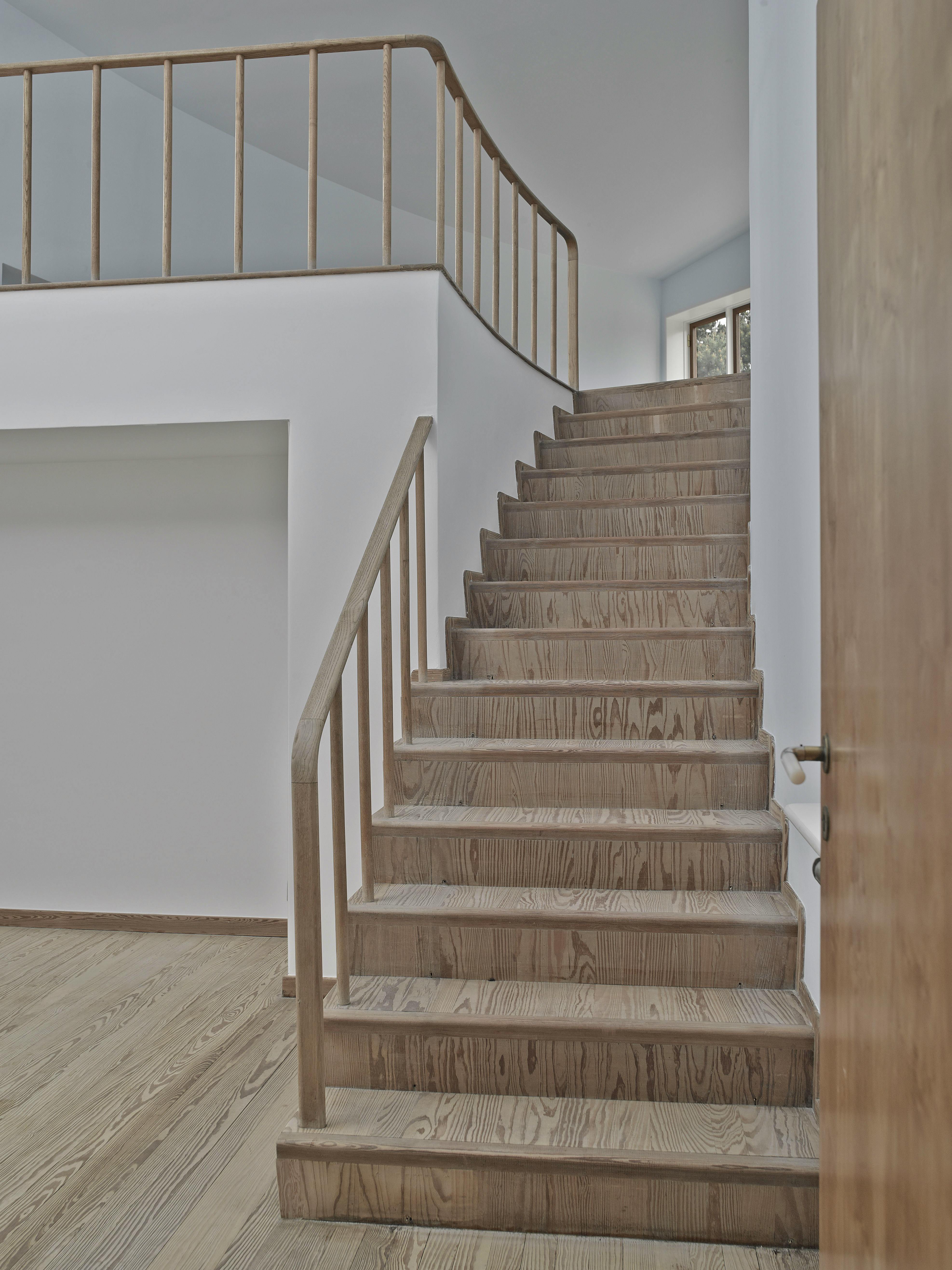 Trætrappe med runde gelænderstolper i to etager i Arne Jacobsens sommerhus. Lyse vægge og trægulv.