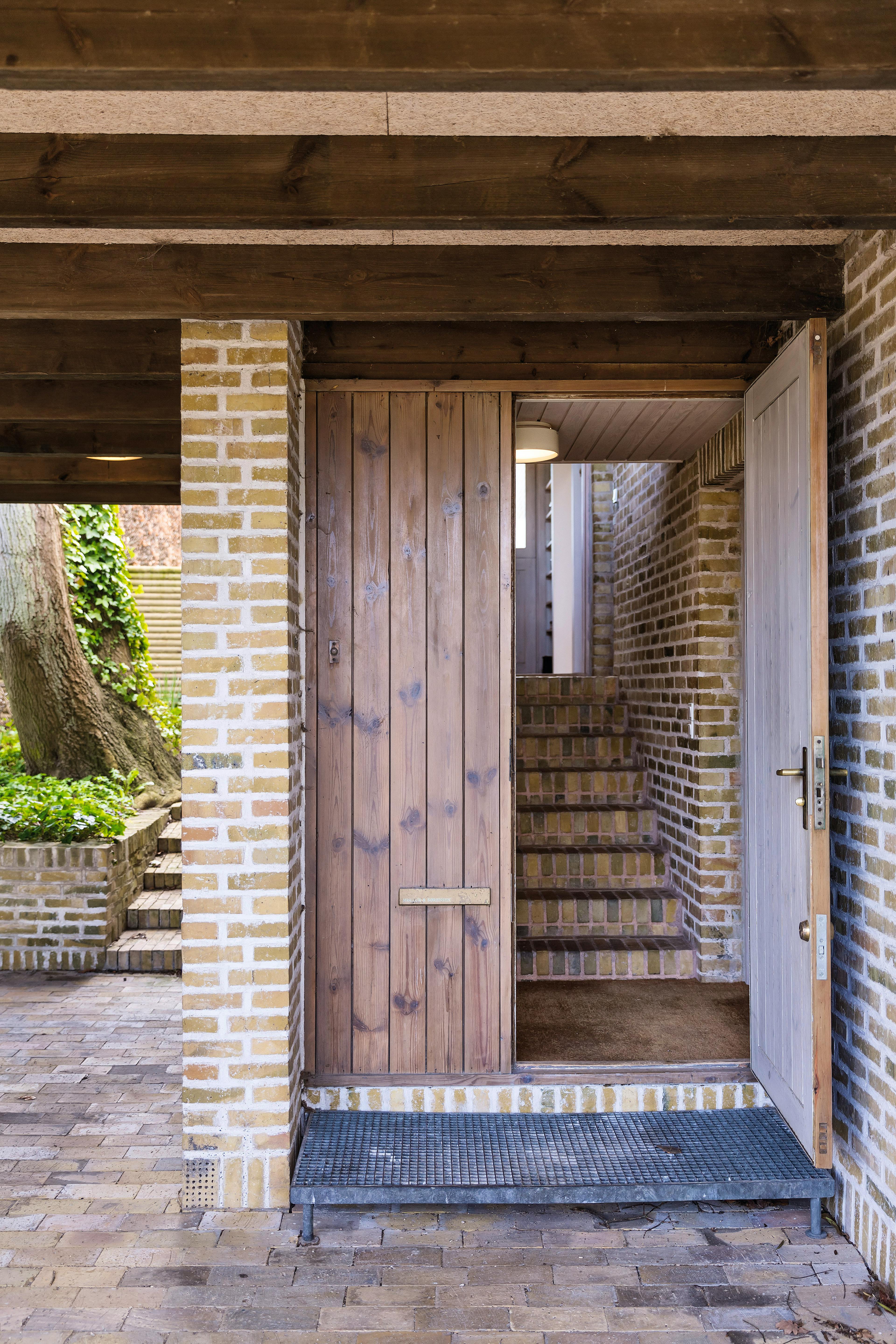 Hoveddøren i Inger og Johannes Exners hus fører ind til en trappegang med gule mursten, omgivet af overdækket murstensgang og træloft.