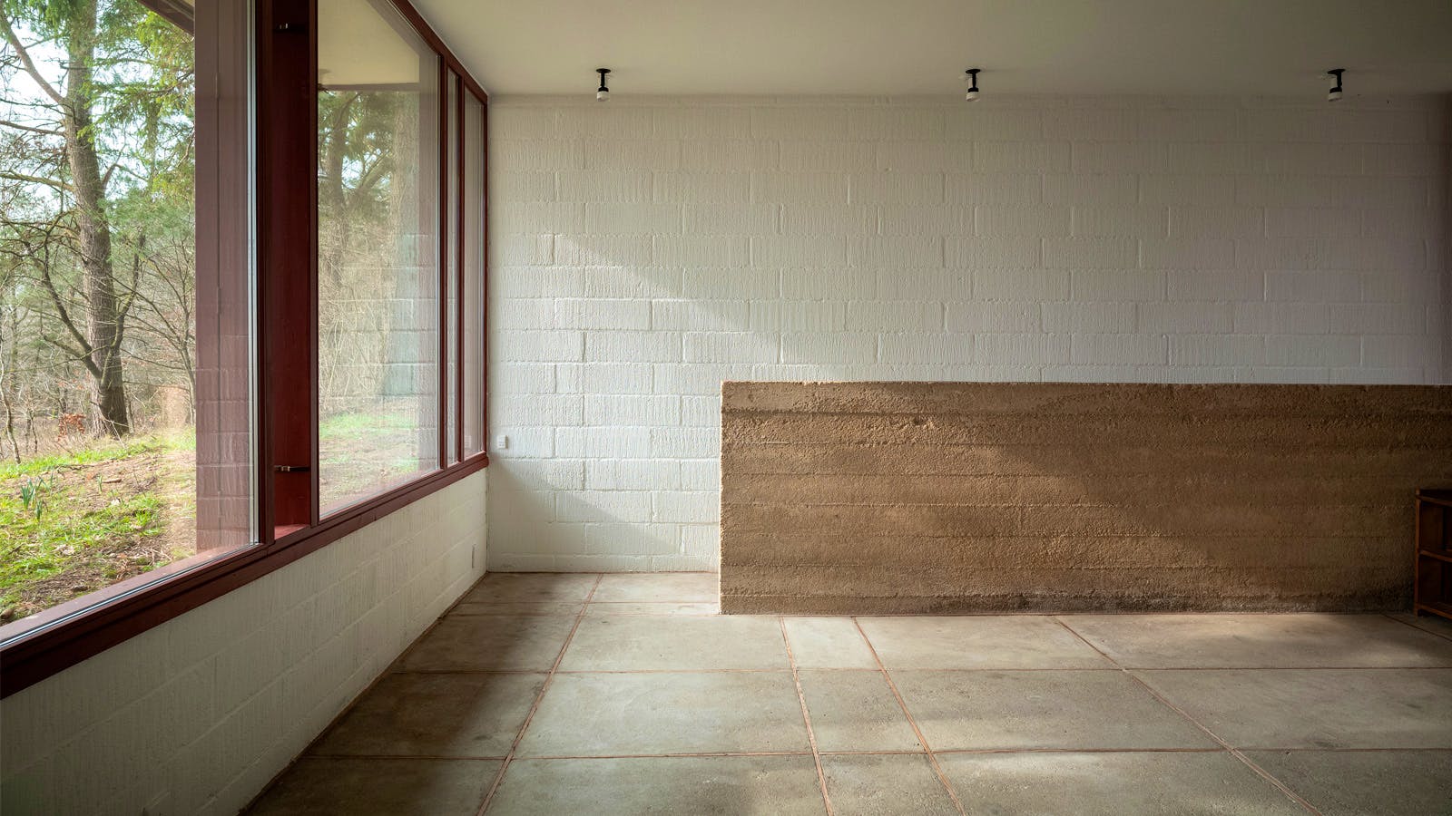 Stue i Poul Erik Thyrrings eget hus med lav halvmur i rå beton, store vinduespartier med rød ramme og gulv af betonfliser.