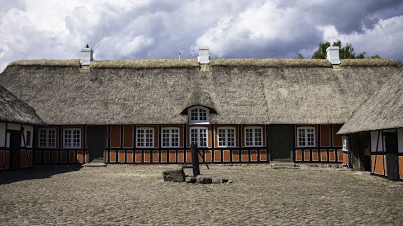 Bindingsværksgård med stråtag og brostensbelagt gårdsplads under dramatiske skyer.