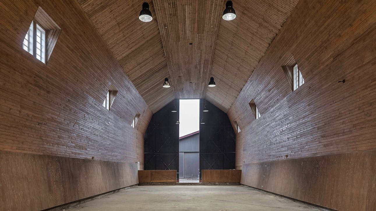 Tom ridehal med højt loft, træbeklædte vægge og små vinduer, oplyst af sorte loftslamper med kig mod portene og udgangen.